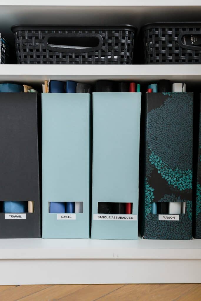 Boîtes de classement étiquetées travail, santé, banque et maison, bien alignées sur une étagère.