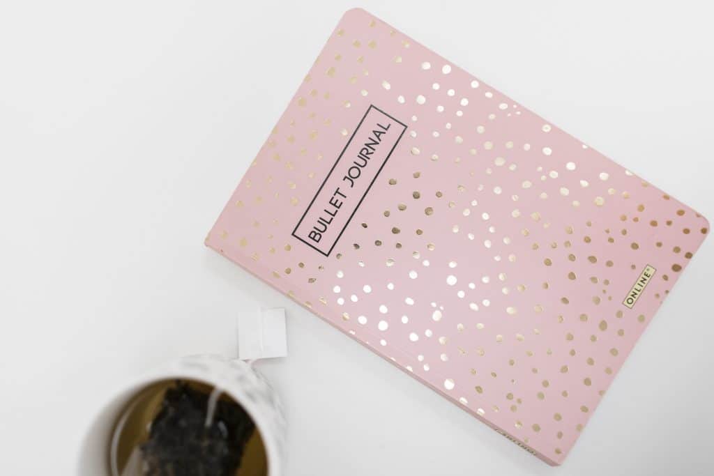 Carnet bullet journal rose doré posé sur une table blanche avec une tasse de thé, symbole d’organisation et de sérénité.